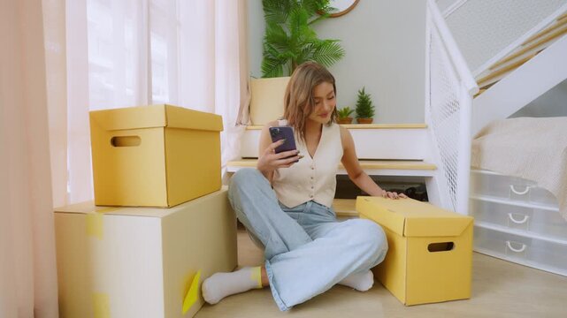 Young Asian woman using mobile smartphone while moving paper box into new house, sitting on stair and chatting to cellphone with her friend