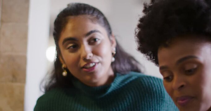 Diverse women leaning viewing offscreen item in kitchen maroon knit speaking green ribbed listening