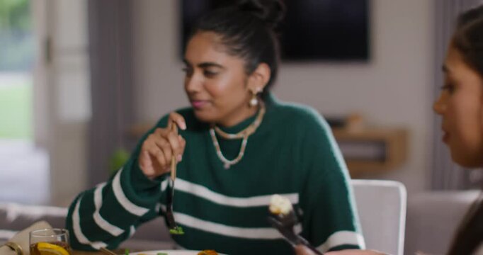 Indian woman in green striped sweater cutting fish with fork, listening to friend speaking at home
