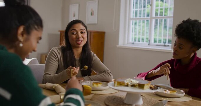 Diverse women friends sitting at table at home, center woman looking left, eating cake with forks