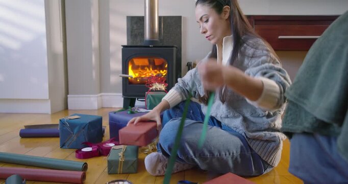 Adult woman sitting cross-legged in sweater tying green ribbons on gifts for holidays by lit stove