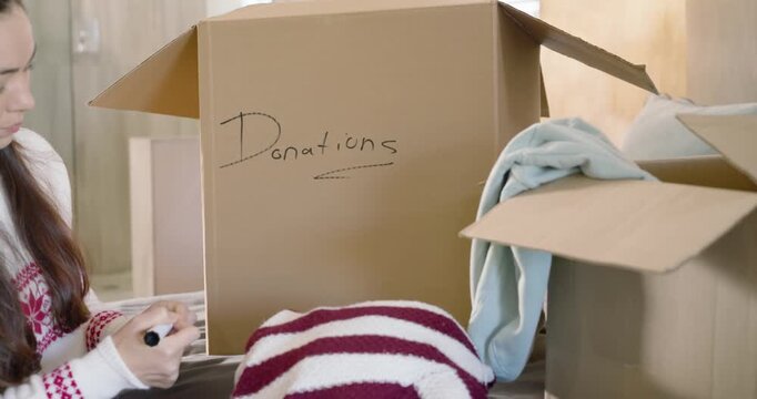 Woman in mid-20s writing Donations label with black marker, picking sweaters and packing box on bed