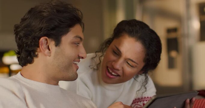 Couple leaning on couch wearing sweaters, watching tablet playing funny clip, reacting and laughing