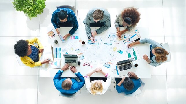 High angle view of a diverse business team collaborating around a white table reviewing reports charts and strategic planning documents during a productive meeting in a bright modern open office works