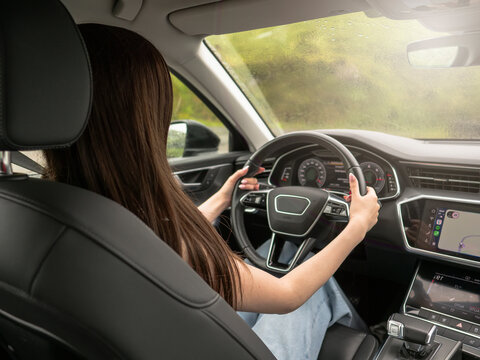 Young woman drives modern car. She looks ahead with calm focus. Hands rest on steering wheel. Car interior shows clean, sleek design. Outside view shows green landscape