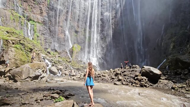 Low angle tilt up video of a young woman looking at the massive majestic cascading flows of Tumpak Sewu waterfall canyon. Epic travel stock footage.