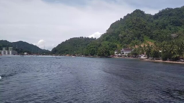 Scenic tropical coastal bay surrounded by lush green forested mountains, coconut palms, small boats, and a peaceful seaside village under a bright cloudy sky. Natural landscape suitable for travel, to