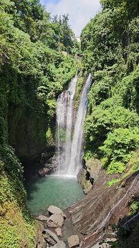 Vertical static shot of Gitgit Waterfall cascading into a turquoise natural pool on Bali island, surrounded by lush green jungle foliage. Nature stock.