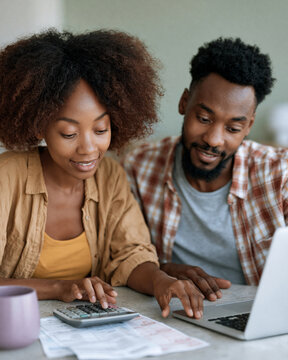 Couple sits at a table, reviewing finances together. Woman smiles gently, hands resting near a calculator. Man looks thoughtful, fingers near laptop keys. They share a quiet moment of planning