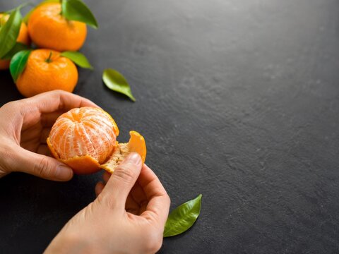 Manos pelando una mandarina sobre una mesa oscura con algunas mandarinas y hojas verdes cerca