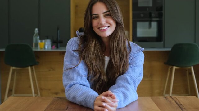 A beautiful young woman with long, dark hair smiles genuinely at the camera from her modern kitchen table. Her warm, friendly expression conveys happiness and positive energy, making for an inviting v