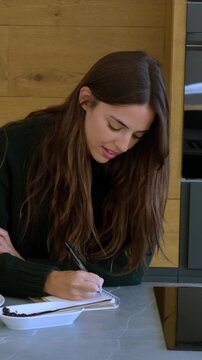 A young, beautiful woman in a green sweater diligently writes in her notebook, taking a thoughtful pause to consider her ideas before continuing her work. This scene captures a moment of focused conce