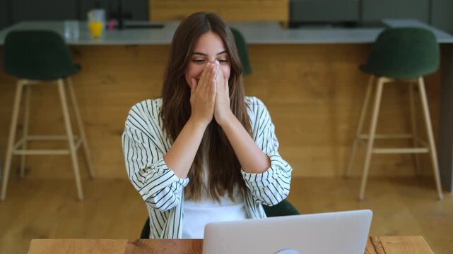 A cheerful young woman celebrates an achievement with immense joy and excitement while working on her laptop. Her happy reaction and successful outcome showcase remote work or online activity in a mod