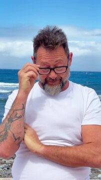 A content and smiling middle-aged man with glasses, a beard, and tattoos stands with arms crossed against a beautiful blue sky and ocean background. He looks up thoughtfully, enjoying the peaceful bea