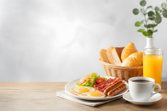 Delicious Full English Breakfast Spread Featuring Fried Eggs, Crispy Bacon, Sausages, Salad, Croissants, Bread Slices, Coffee, and Orange Juice on a Wooden Table with Copy Space for Text