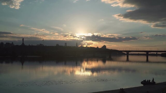 Sunset light breaks through clouds over a wide river Dnieper as a Kyiv city skyline and bridge form dark silhouettes, with calm reflections and a small boat drifting near the shore