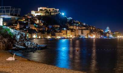 Naklejka premium Night view of Sibenik historic old town with illuminated fortress, cathedral, and a white swan on the pebble beach