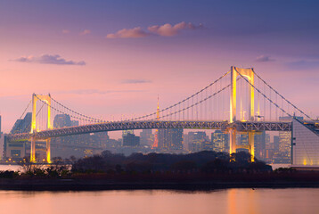 Obraz premium Rainbow Bridge illuminated at twilight with Tokyo skyline and Tokyo Tower in the background, reflecting on calm waterfront under a colorful pastel sunset sky in Tokyo, Japan.