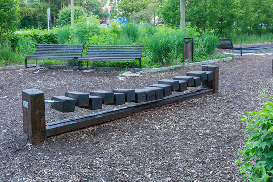 Low wooden balance obstacle made from suspended square blocks stands on a woodchip surface beside benches, ornamental grasses and a litter bin. The outdoor installation supports balance training, coor