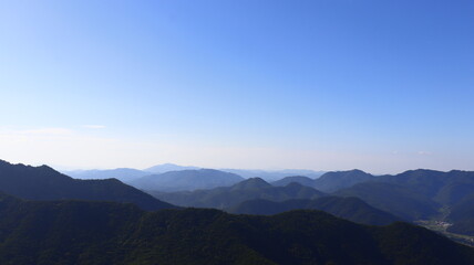Fototapeta premium Expansive View of Layered Mountains and Forest with Distant Valley Village Under Clear Blue Sky
