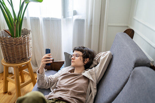 Bored woman relaxing on couch at home using smartphone, browsing social media procrastinating time, laziness and idling female comfortably lying on sofa checking news feeds online on mobile.
