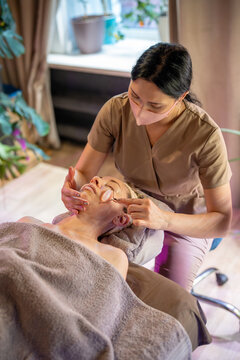 Beautician in spa clinic making stone roller face massage for female client lying on spa table, using tool with polished stones, gliding on skin to increase circulation and reduce facial puffiness.