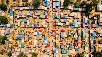 Naklejka premium Aerial view of a bustling outdoor market featuring a vibrant grid of colorful vendor stalls with people walking through narrow sandy pathways under the bright warm morning sun in Africa.