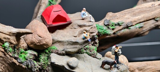Miniature camping diorama featuring a small red tent on rocky terrain surrounded by figurines and natural elements. Driftwood, moss, and stones enhance the realistic outdoor setting. © Johari