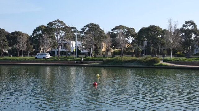 Waterfront view of the man-made lake at Sanctuary Lakes estate in Point Cook, Melbourne Australia. Calm water with colorful floating buoys, lined by contemporary residential suburban houses