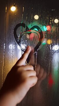 A child finger traces a heart onto a fogged-up windowpane during a rainy night. Through the condensation and dripping water, the colorful, out-of-focus city lights create a warm, glowing bokeh