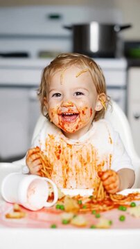 A joyful toddler sits in a high chair, covered head-to-toe in bright red spaghetti sauce. Clutching noodles in tiny hands, the child beams with a messy, toothy grin. Hilarious first self-fed meal