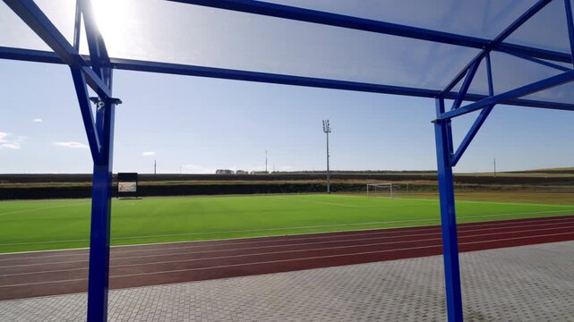 Bright football pitch and running tracks extend behind metal canopy framework. Clean empty outdoor arena remains quiet under clear sky