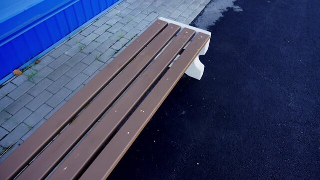 Slatted bench with concrete legs stands between asphalt pathway and paving stones. Clean outdoor seating area offers quiet resting spot
