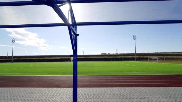Wide soccer field and running track stretch under metal canopy structure. Empty outdoor stadium looks peaceful under clear afternoon sky