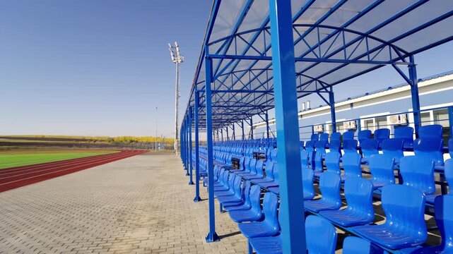 Plastic seats stand under canopy near outdoor running track. Empty spectator stands remain quiet under sunny sky before competition begins