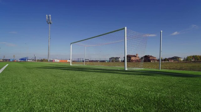 Football goal stands on green grass pitch next to painted line. Tall lighting tower under blue sky creates professional sports atmosphere