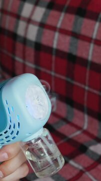 A light blue toy bubble gun blows iridescent soap bubbles. A hand holds the plastic device against a soft red plaid blanket. The colorful bubbles float gently through the air.
