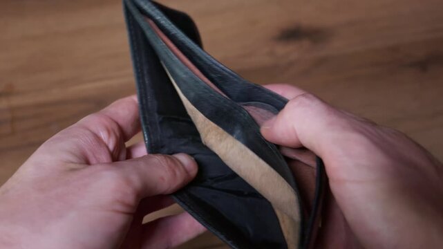 Close-up view of a person hands opening an empty black leather wallet to reveal no money inside. The camera captures the interior lining against a blurred wooden table background.