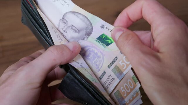 Hands open a black leather wallet filled with five hundred Ukrainian Hryvnia banknotes on a wooden table. Concept of personal finance, cash savings, wealth, and economy in Ukraine.