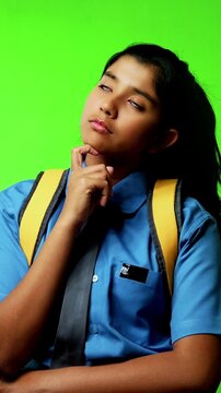 Portrait of Indian Asian teenage girl in school uniform with backpack standing against green chroma screen, looking thoughtful while placing hand on her chin, symbolizing curiosity, decision-making