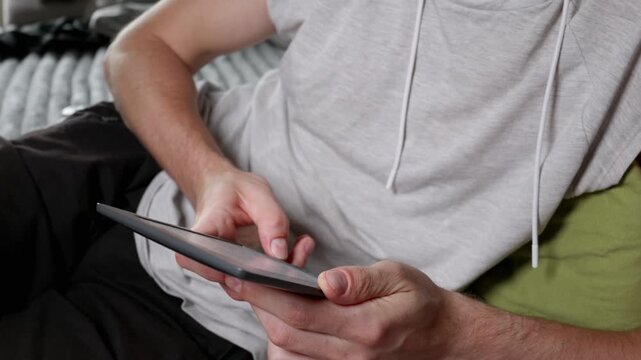 Man uses his thumbs to scroll through text on a sleek grey tablet while sitting in a relaxed position. Concept of digital literacy, modern education, leisure time, and home entertainment.