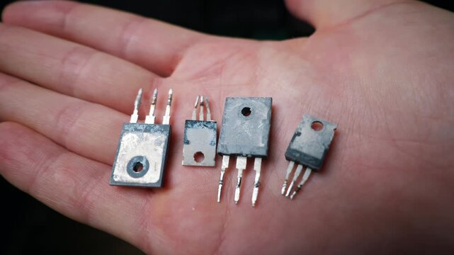 A close-up view of four used electronic transistors resting on a person open palm. These semiconductors show white thermal paste residue and soldered pins. Soft lighting highlights the details.