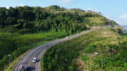 Obraz premium Drone shot of cars driving on a curvy highway along steep cliffs and tropical forests on a sunny day.