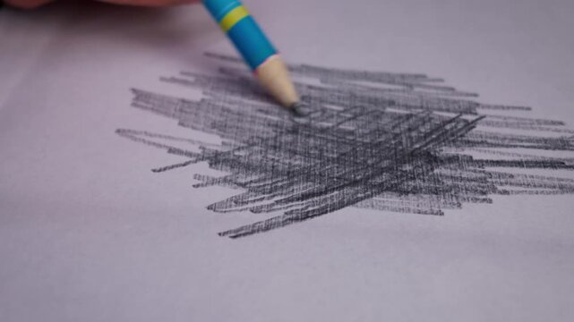 A hand holds a blue and yellow pencil while scribbling dense graphite lines onto a sheet of white paper. The artist moves the pencil rapidly to create a dark patch of overlapping strokes.