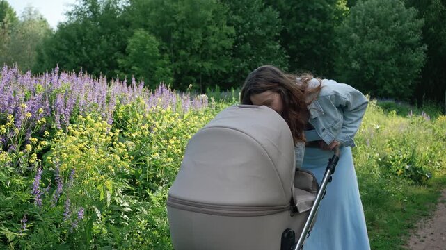 Young loving mother walking with a baby stroller in a lush green park on a sunny day, stopping to look at her newborn child amidst a field of blooming purple flowers