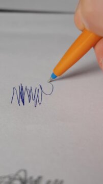 A macro view of a person writing on white paper with a bright orange ballpoint pen. The blue ink smoothly glides onto the clean page under bright and natural morning light.