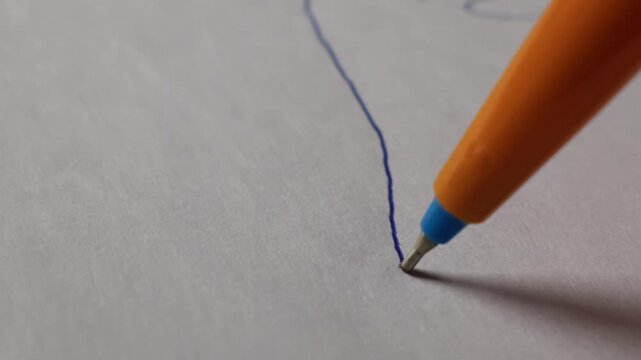 Orange ballpoint pen creates a thin blue ink line on a white textured paper surface during a close-up macro shot. Concept of creative writing, academic study, and artistic sketching process.