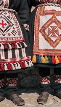Traditional folk clothing with embroidered aprons geometric red white black orange patterns tassels fringes wool skirts knitted socks leather shoes cultural textile heritage generated ai