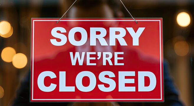 Red "Sorry We're Closed" sign hanging on glass door, with blurry lights