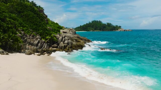 Turquoise waves gently hitting a white sand beach surrounded by lush green vegetation and rocky formations. Seychelles, Mahe.